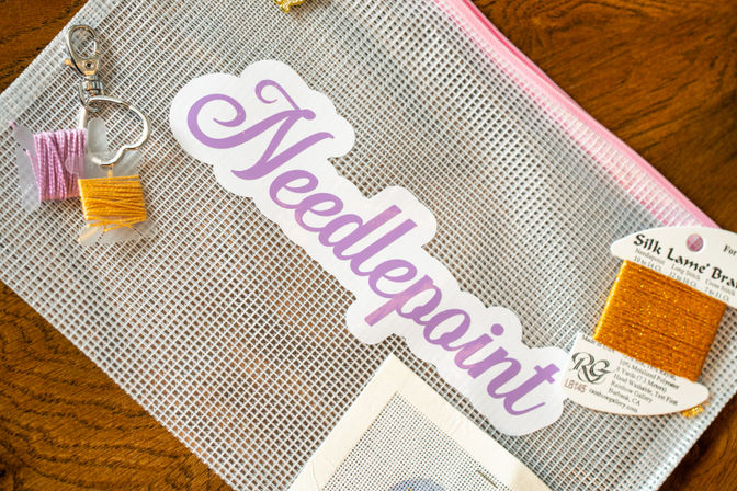 Purple "Needlepoint" sticker on a white mesh craft pouch with a pink zipper, surrounded by gold and yellow embroidery threads and a small pink-and-yellow bobbin keychain on a wooden table.