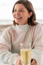 Smiling young woman in a pastel quilted floral sweater and pearl earring, holding a blurred champagne glass at a beachside table with the ocean in the background