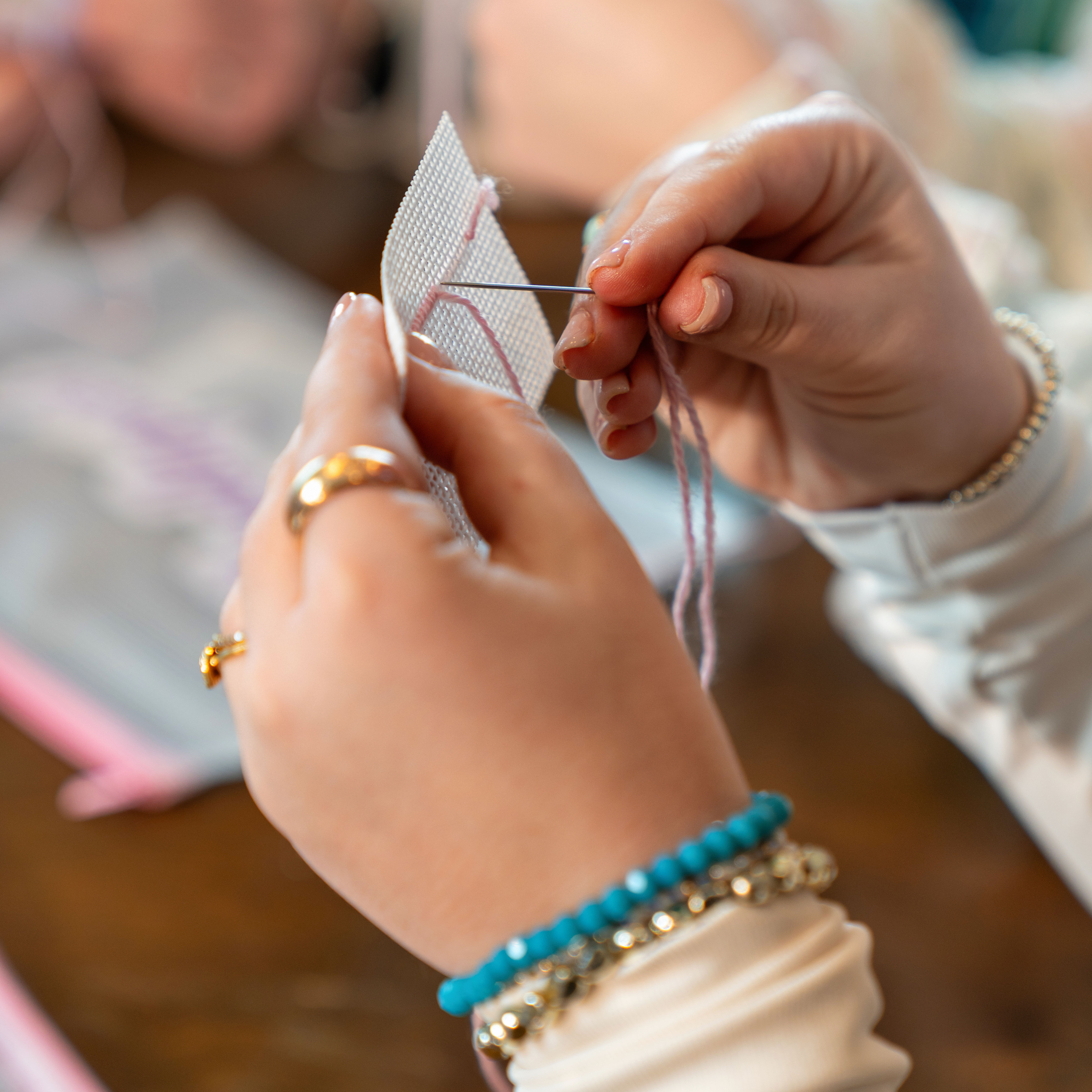Close-up of hands stitching pink cross‑stitch on white Aida cloth with a threaded needle, turquoise beaded bracelet and gold rings visible in a cozy indoor craft scene.