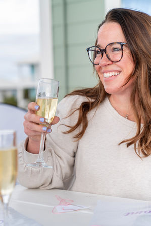 Smiling woman wearing glasses toasting with a champagne flute on a sunny coastal patio, casual sweater and pastel nail polish.