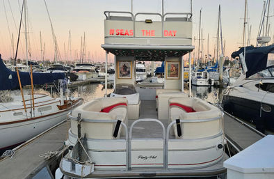 Pontoon boat docked in a marina at sunset, cushioned seating and “#SEAS THE DAY” sign on the upper deck, surrounded by sailboats and tall masts reflecting on calm water.