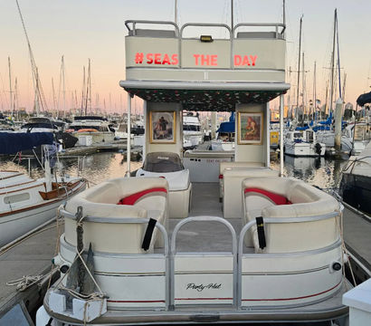 Pontoon boat docked in a marina at sunset, cushioned seating and “#SEAS THE DAY” sign on the upper deck, surrounded by sailboats and tall masts reflecting on calm water.