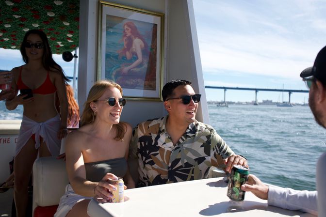 Friends laughing on a sunny boat ride, holding canned drinks at a small table with a framed mermaid print, bay waters and a long coastal bridge in the background.