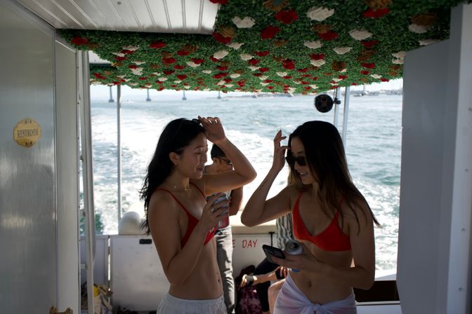 Two women in red bikinis laughing on a floral-canopied boat deck, holding canned drinks and a phone with sunlit ocean and bridge supports in the background.