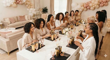 Cheerful group of women in coordinating robes toasting with champagne at a marble kitchen island, surrounded by blush balloon garland and floral centerpieces in a chic home celebration.