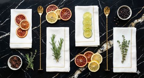 Flat-lay of cocktail garnishes on black marble — white linen napkins with rosemary and thyme, lime and blood orange slices, star anise, juniper berries and twisted gold bar spoons.