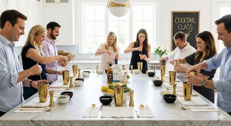 Group of adults taking a cocktail class around a marble kitchen island in a bright white modern kitchen, stirring and pouring drinks into gold shakers with bottles, citrus bowls and a 'Cocktail Class' sign in the background.