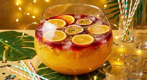 Fishbowl-style glass punch bowl with layered tropical punch, orange slices and passion fruit halves over ice, surrounded by colorful paper straws, confetti and warm fairy lights on a festive table.