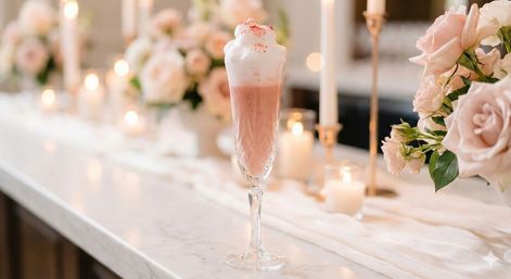 Pink frothy cocktail in a crystal champagne flute on a marble countertop, surrounded by blush roses, flickering candles and a soft romantic tablescape.