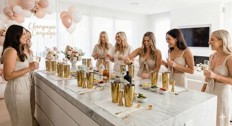 Sunlit modern kitchen party with a group of women in neutral linen outfits gathered around a marble island mixing cocktails and holding champagne flutes, gold shakers and barware, pink and white balloons and floral arrangements for a celebratory brunch