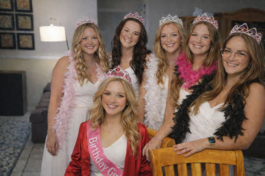 Smiling group of women in white dresses and tiaras celebrating a birthday in a cozy living room; the birthday girl sits front wearing a pink sash and red robe while friends pose with feather boas.