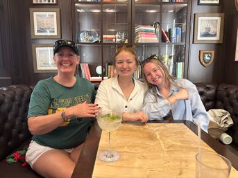 Three smiling people seated at a marble-top table in a cozy nautical-themed lounge, tufted leather banquette and glass-front bookshelves behind them, one holding a large gin-and-tonic with lime.