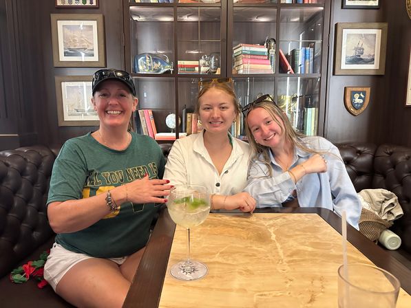 Three smiling people seated at a marble-top table in a cozy nautical-themed lounge, tufted leather banquette and glass-front bookshelves behind them, one holding a large gin-and-tonic with lime.