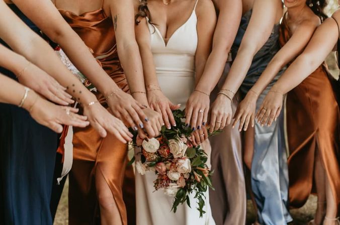 Bride in white holding a floral bouquet as bridesmaids in satin copper, navy and silver dresses reach hands forward at an outdoor wedding