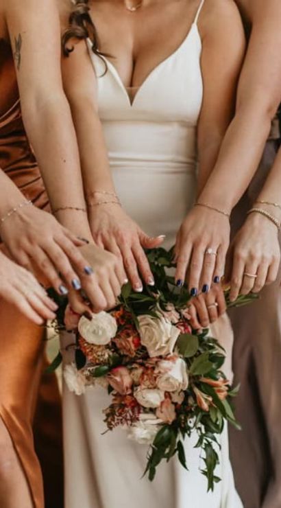Bride in white holding a floral bouquet as bridesmaids in satin copper, navy and silver dresses reach hands forward at an outdoor wedding