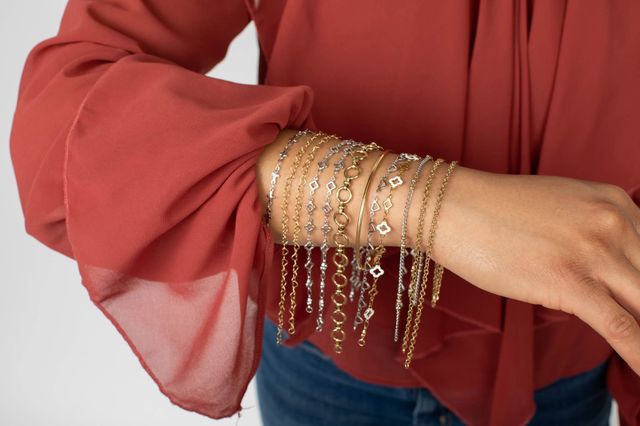 Layered gold and silver chain bracelets cascading over a person's wrist, styled with a flowing rust-red blouse sleeve — close-up fashion accessory shot.