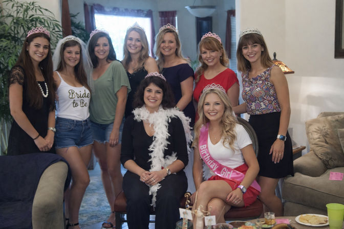 Smiling group of women in a living room wearing tiaras at a combined bachelorette and birthday party — one in a veil and another wearing a pink "Birthday Girl" sash.
