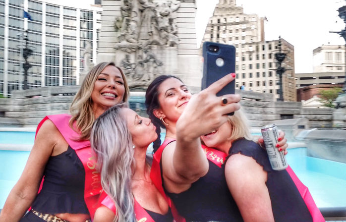 Four women wearing pink sashes smiling and taking a selfie by a downtown fountain and historic monument with city skyline in the background