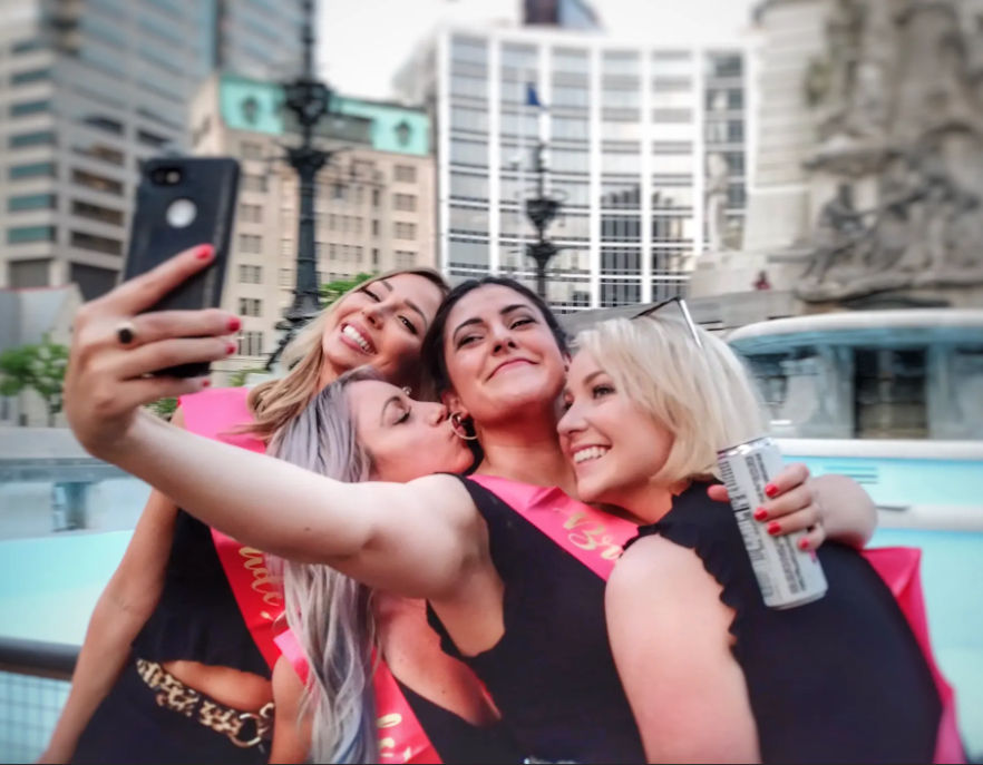 Four friends wearing pink celebration sashes take a joyful selfie in a downtown city plaza by a fountain and modern office buildings; one holds a canned drink while another kisses a cheek.