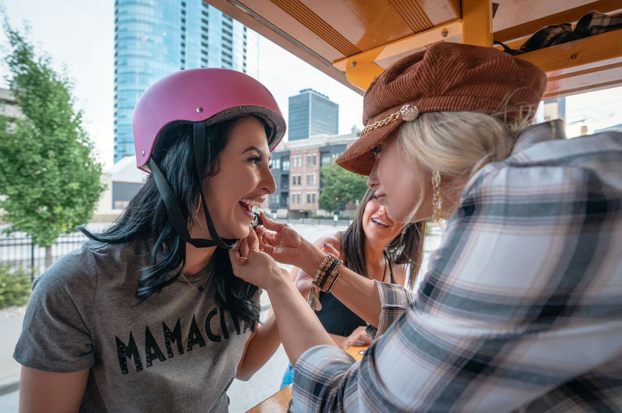 Three friends laughing on a downtown pedal bike; woman in a pink helmet and ‘MAMACITA’ tee gets lipstick applied by a friend with city skyline in the background.