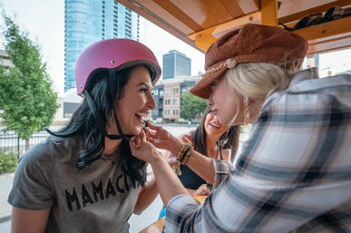 Three friends laughing on a downtown pedal bike; woman in a pink helmet and ‘MAMACITA’ tee gets lipstick applied by a friend with city skyline in the background.