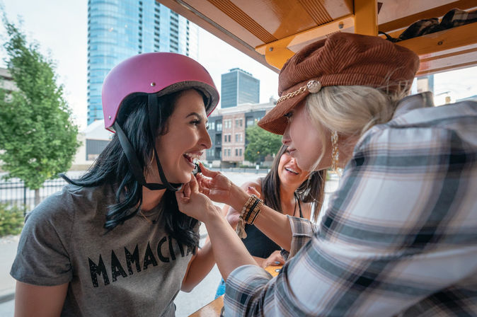 Three friends laughing on a downtown pedal bike; woman in a pink helmet and ‘MAMACITA’ tee gets lipstick applied by a friend with city skyline in the background.