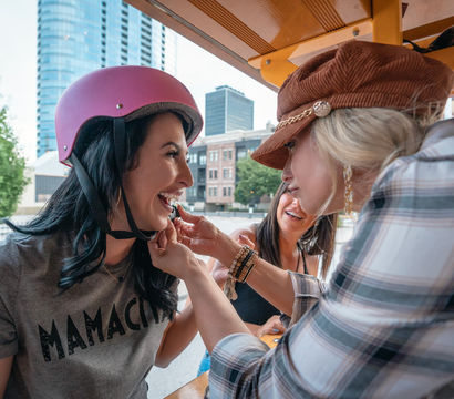 Three friends laughing on a downtown pedal bike; woman in a pink helmet and ‘MAMACITA’ tee gets lipstick applied by a friend with city skyline in the background.