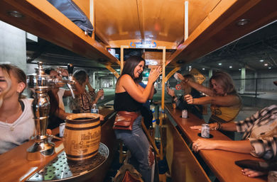 Friends laughing and clinking drinks aboard a pedal-powered mobile bar in a city parking garage, with beer taps and a wooden keg on the counter.