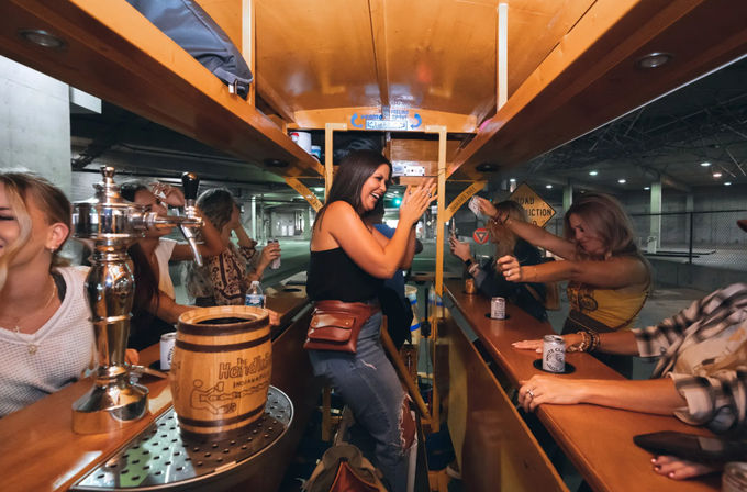 Friends laughing and clinking drinks aboard a pedal-powered mobile bar in a city parking garage, with beer taps and a wooden keg on the counter.