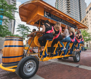 Group of women wearing pink sashes pedaling a bright yellow pedal-bar party bike past high-rise buildings on a downtown urban street with a wooden barrel at the front.