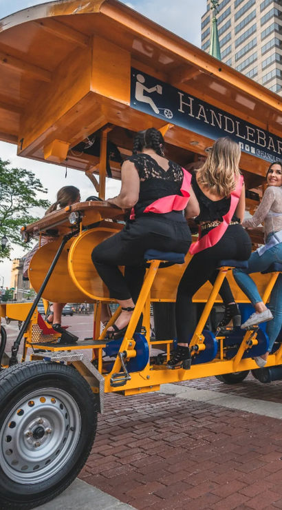 Group of women wearing pink sashes pedaling a bright yellow pedal-bar party bike past high-rise buildings on a downtown urban street with a wooden barrel at the front.