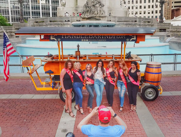 Seven women wearing bachelorette sashes pose in front of a bright orange pedal bar with an American flag at the monument fountain in downtown Indianapolis.