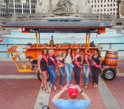 Seven women wearing bachelorette sashes pose in front of a bright orange pedal bar with an American flag at the monument fountain in downtown Indianapolis.