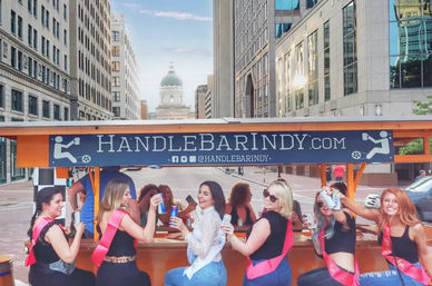 Group of friends on a pedal-powered party bike in downtown Indianapolis, women wearing sashes laughing and toasting canned drinks with a historic domed building and city skyscrapers in the background.
