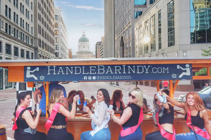 Group of friends on a pedal-powered party bike in downtown Indianapolis, women wearing sashes laughing and toasting canned drinks with a historic domed building and city skyscrapers in the background.