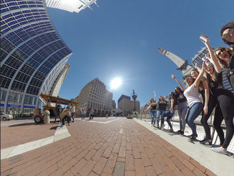 Wide-angle street view of a group of friends celebrating on the sidewalk at Monument Circle in downtown Indianapolis, with the Soldiers and Sailors Monument, modern glass buildings, a mobile pedal bar, and a bright sunny sky.
