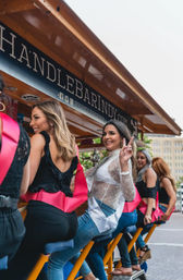 Smiling women in sashes riding a colorful pedal-party bike on a downtown street, one flashing a peace sign.