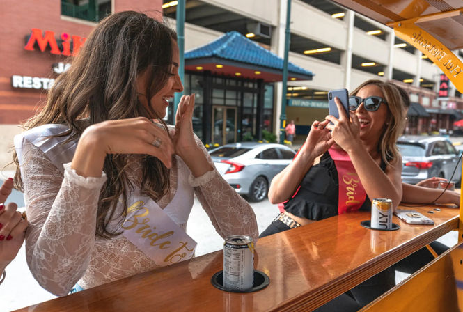 Two women wearing bride-to-be sashes laugh at a wooden party-bar counter on a downtown street, one snapping a photo with her phone while canned drinks sit in cup holders.
