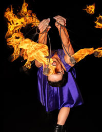 Fire performer in a purple costume spinning flaming poi, bright orange flames arcing midair against a black background in a dramatic night stage show.