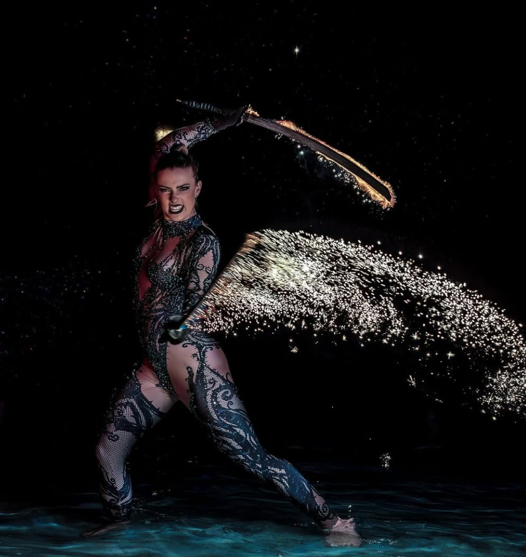 Night fire performer in ornate bodysuit swings a spark-emitting whip over shallow water, showering glittering pyrotechnic sparks in a dramatic live show