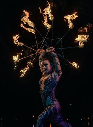 Fire dancer in a sparkling beaded bodysuit spins a multi-spoke fire fan, flames forming a dramatic halo against a dark night backdrop