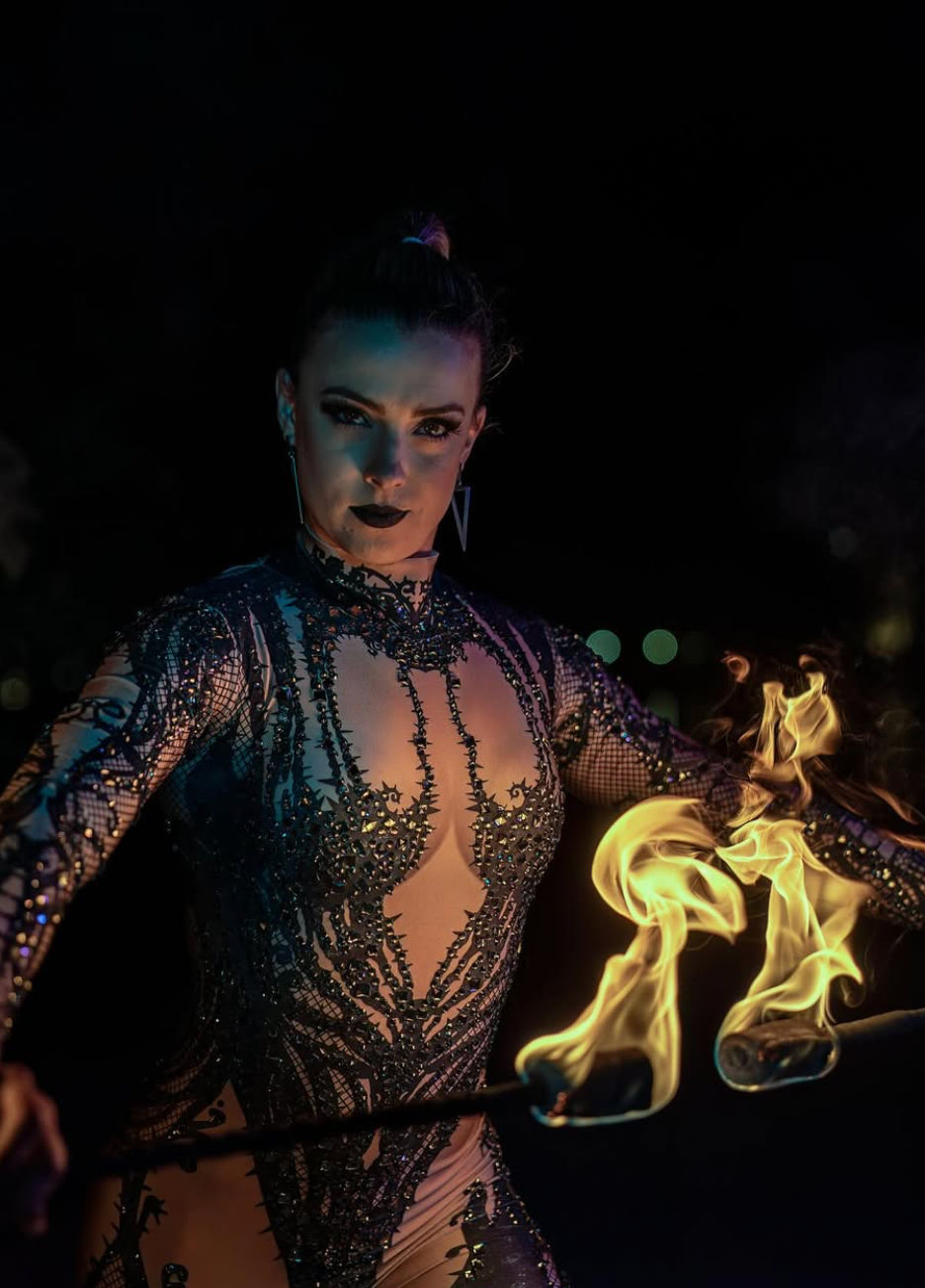 Fire performer in an ornate black beaded bodysuit and dramatic makeup holding two flaming hand torches against a dark night background.