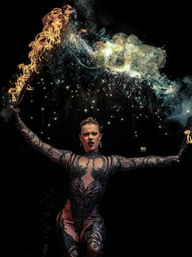 Fire performer in ornate black bodysuit holding flaming torches overhead, creating an arc of fire, smoke and sparkling embers against a dark stage — dramatic circus-style fire show.
