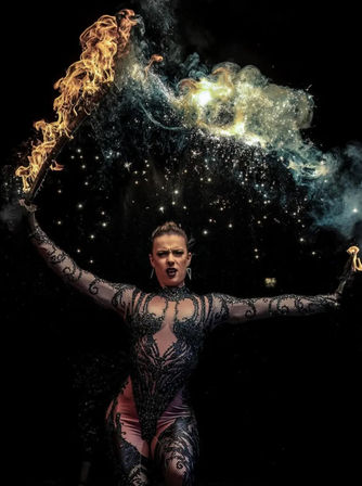 Fire performer in ornate black bodysuit holding flaming torches overhead, creating an arc of fire, smoke and sparkling embers against a dark stage — dramatic circus-style fire show.