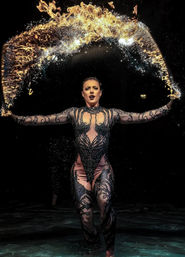 Fire performer in an intricate black bodysuit tossing a shimmering arc of flame and water across a dark stage.