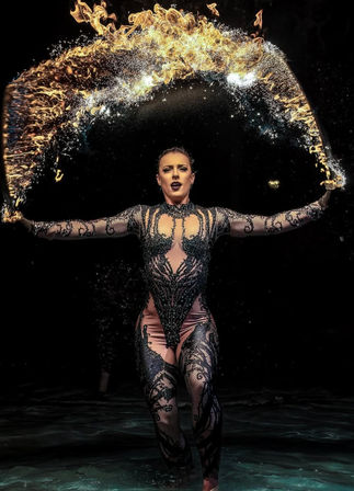 Fire performer in an intricate black bodysuit tossing a shimmering arc of flame and water across a dark stage.