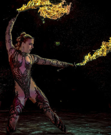 Fire performer twirling two flaming staffs in a dramatic pose, wearing an ornate mesh bodysuit as yellow flames and sparks light a dark stage backdrop.