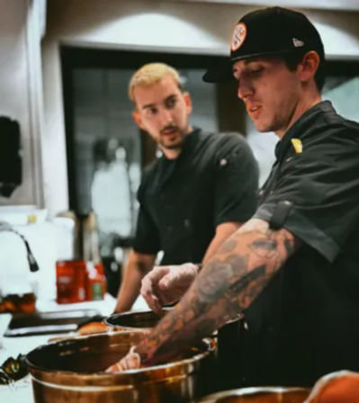 Two chefs collaborating in a restaurant kitchen: a tattooed chef wearing a cap stirs large metal mixing bowls while another chef watches and consults.