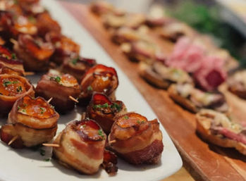 Bite-sized bacon-wrapped morsels glazed and sprinkled with herbs on a white platter, toothpicked for serving, with assorted crostini on a wooden board in the blurred background — party appetizer spread.