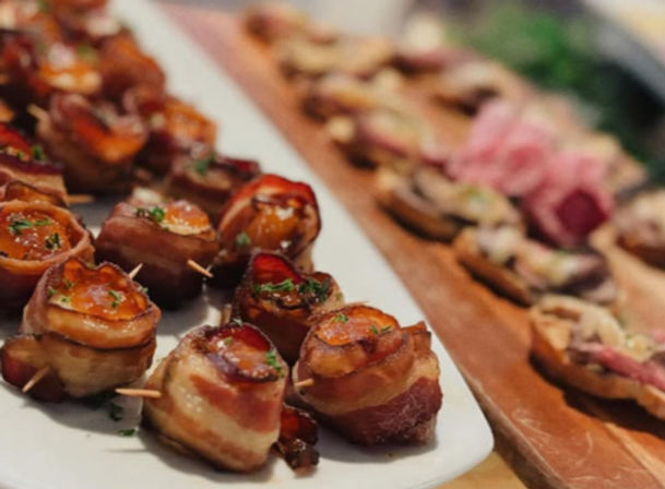 Bite-sized bacon-wrapped morsels glazed and sprinkled with herbs on a white platter, toothpicked for serving, with assorted crostini on a wooden board in the blurred background — party appetizer spread.